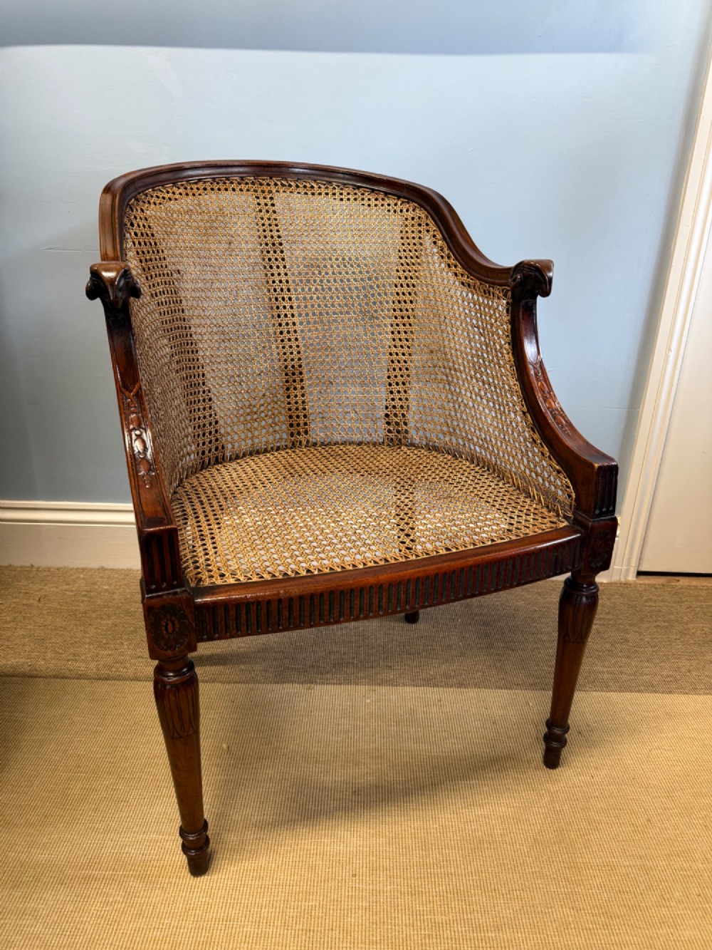 An antique mahogany bergere armchair featuring hand-woven cane panels, a curved backrest, fluted seat rail, and turned front legs.