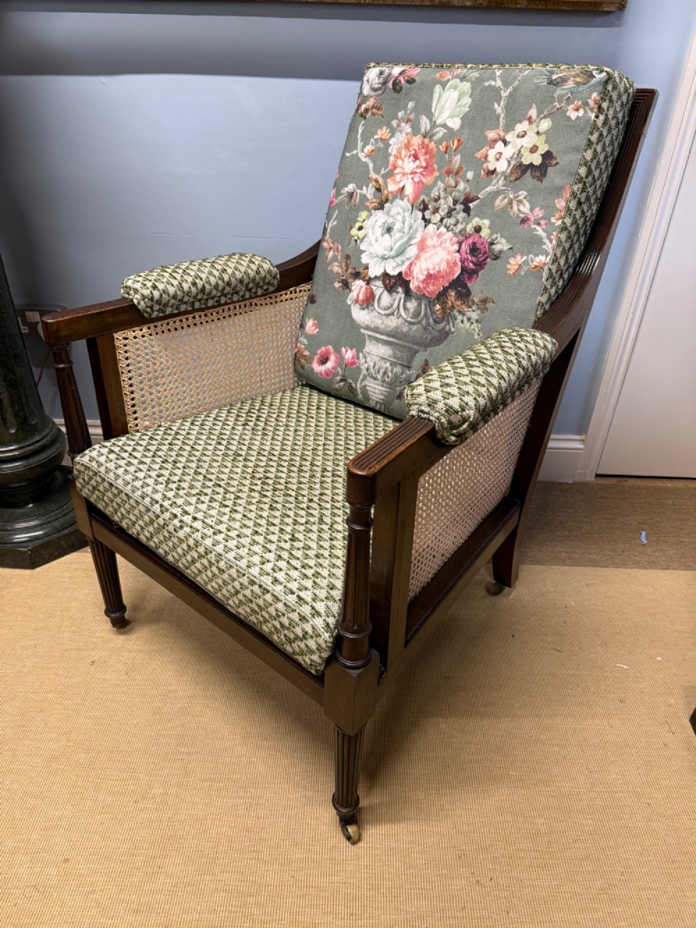 A late 19th-century mahogany bergere armchair with woven cane sides, fluted front legs on brass castors, green patterned seat cushion, and a floral upholstered backrest.