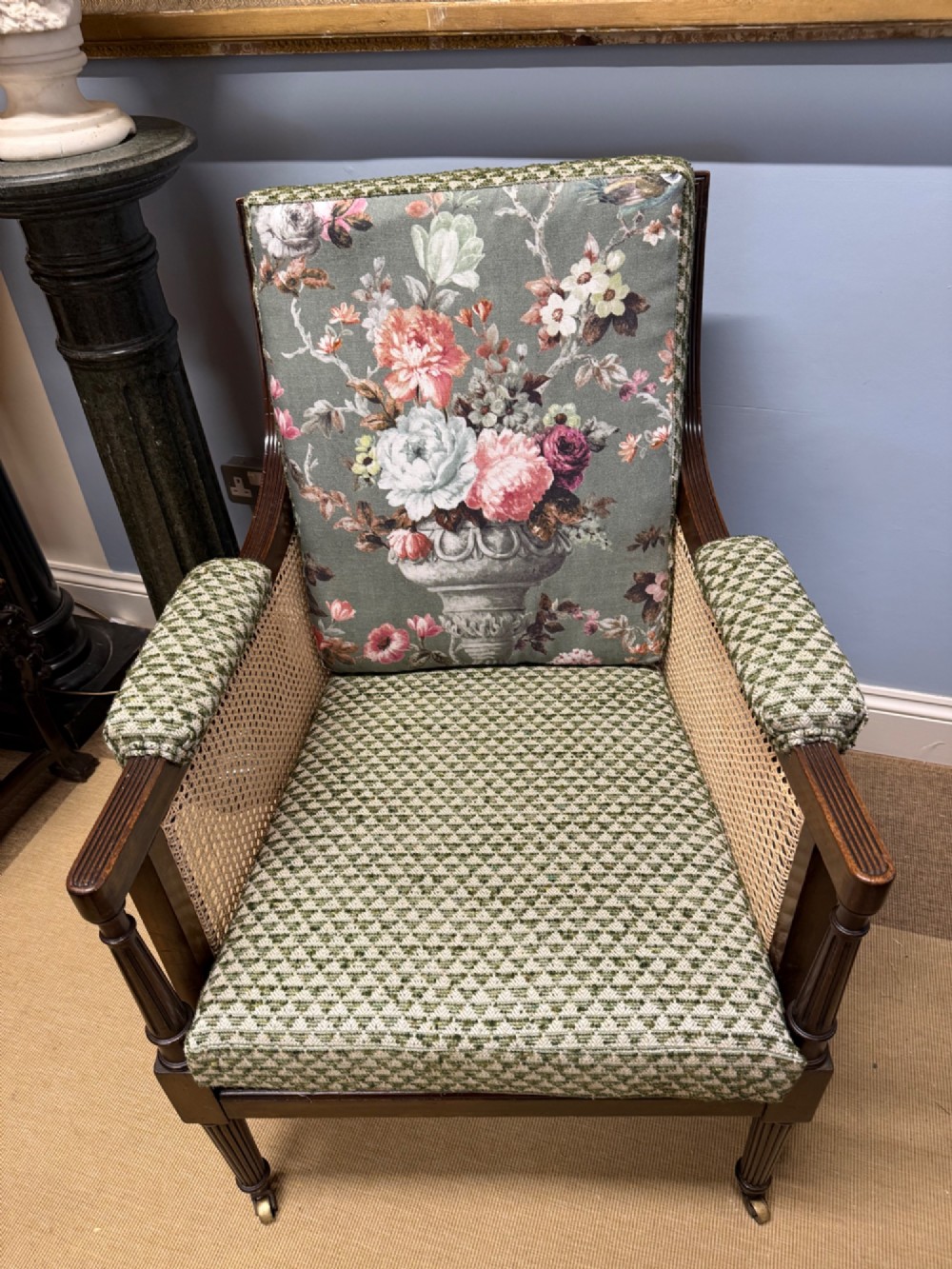 A late 19th-century mahogany bergere armchair with woven cane sides, fluted front legs on brass castors, green patterned seat cushion, and a floral upholstered backrest. View III