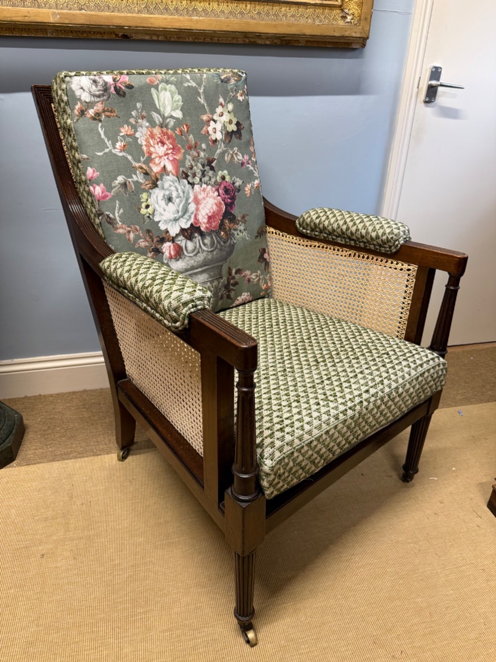 A late 19th-century mahogany bergere armchair with woven cane sides, fluted front legs on brass castors, green patterned seat cushion, and a floral upholstered backrest. View V