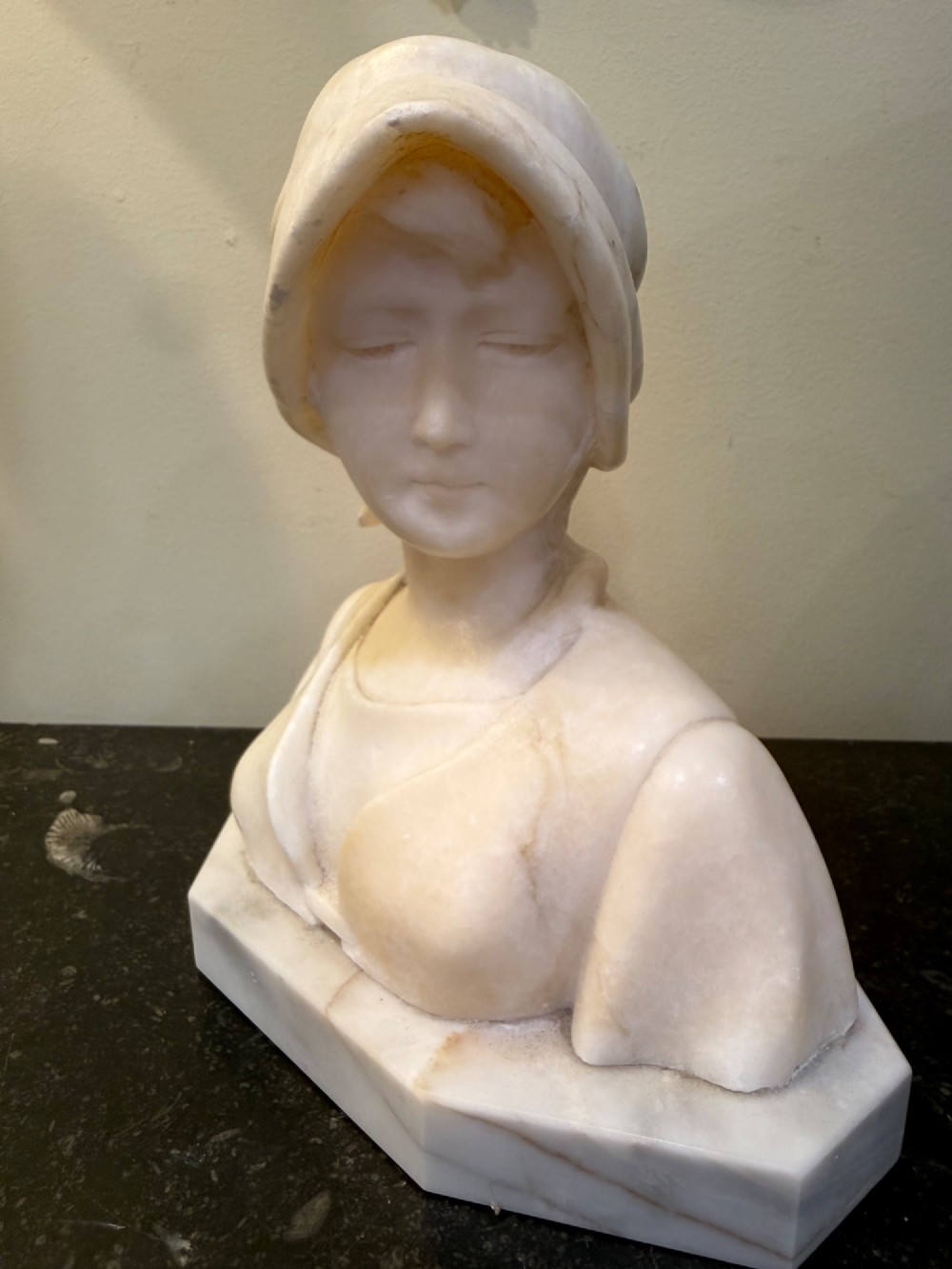An off-white carved alabaster bust depicting a young woman wearing a bonnet, resting on an integral octagonal stone base.