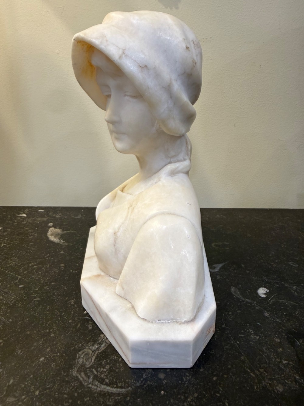 An off-white carved alabaster bust depicting a young woman wearing a bonnet, resting on an integral octagonal stone base. View II