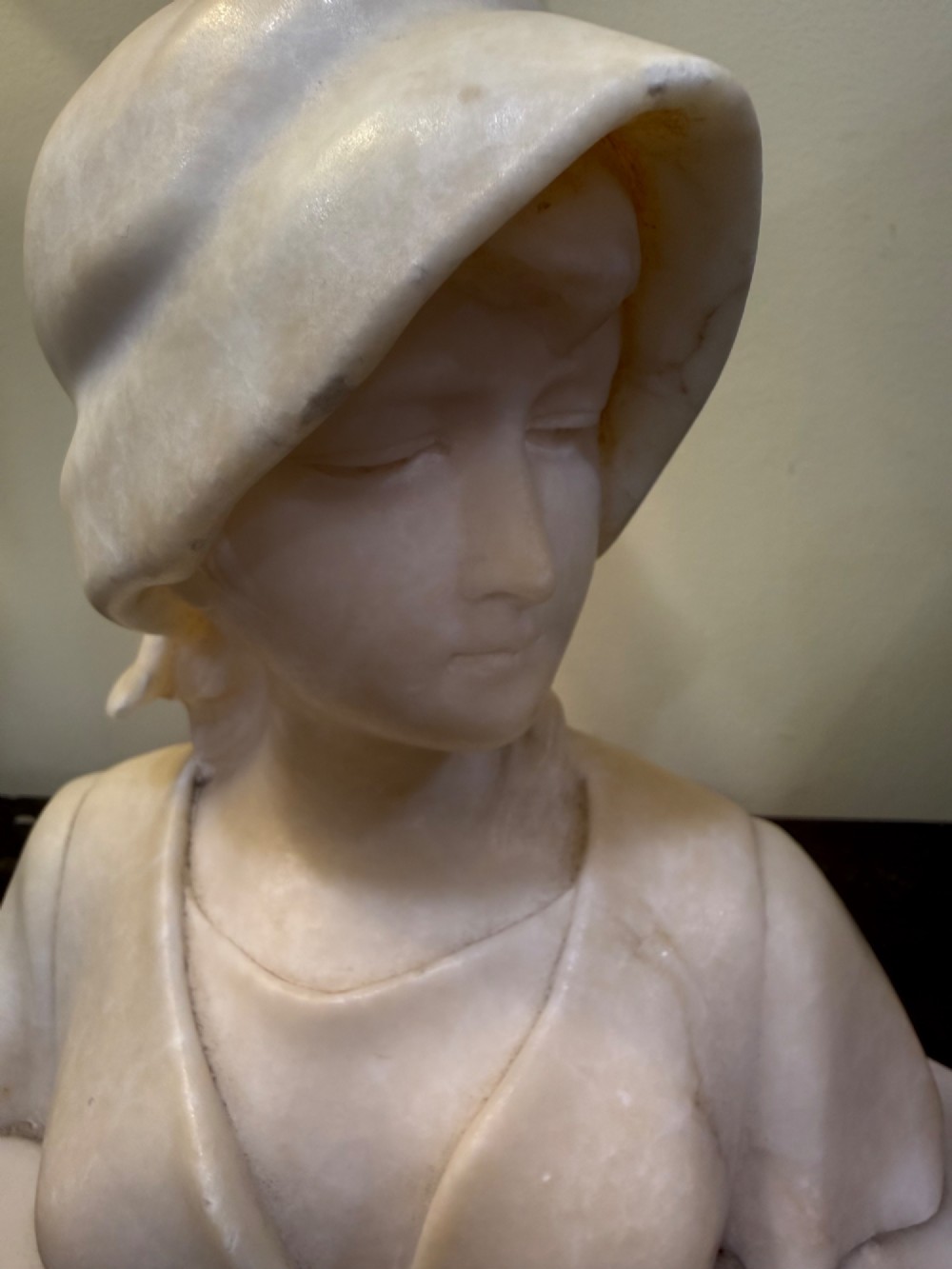 An off-white carved alabaster bust depicting a young woman wearing a bonnet, resting on an integral octagonal stone base. View V