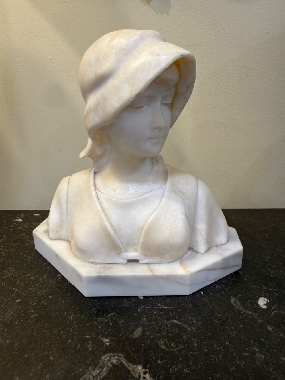An off-white carved alabaster bust depicting a young woman wearing a bonnet, resting on an integral octagonal stone base. View VI