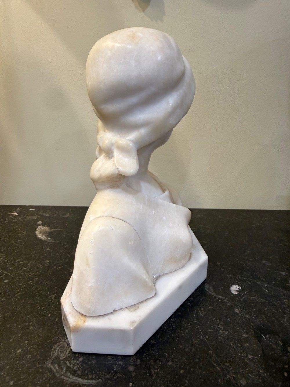 An off-white carved alabaster bust depicting a young woman wearing a bonnet, resting on an integral octagonal stone base. View VII