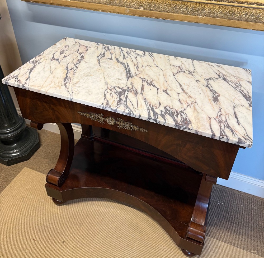 A 19th-century mahogany console table featuring a white veined marble top, an ornate brass mount on the apron, curved scroll front supports, a plinth base, and a mirrored back. View II