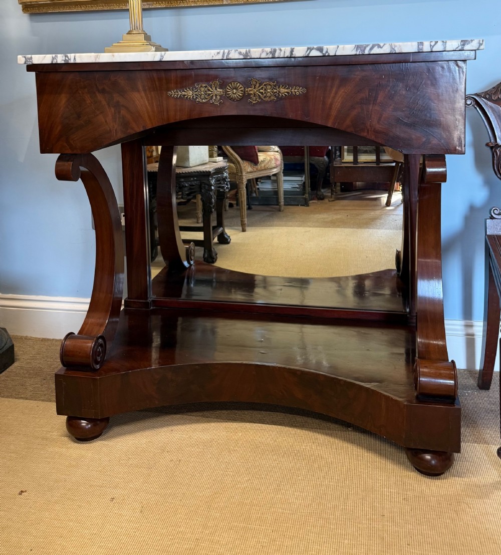 A 19th-century mahogany console table featuring a white veined marble top, an ornate brass mount on the apron, curved scroll front supports, a plinth base, and a mirrored back.