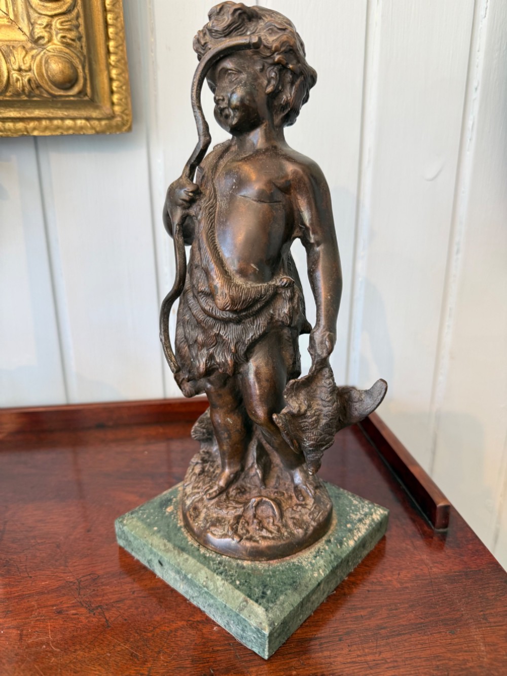 An antique dark bronze figural sculpture depicting a young cherub holding a bow and hunted game, standing on a square green marble base. View II