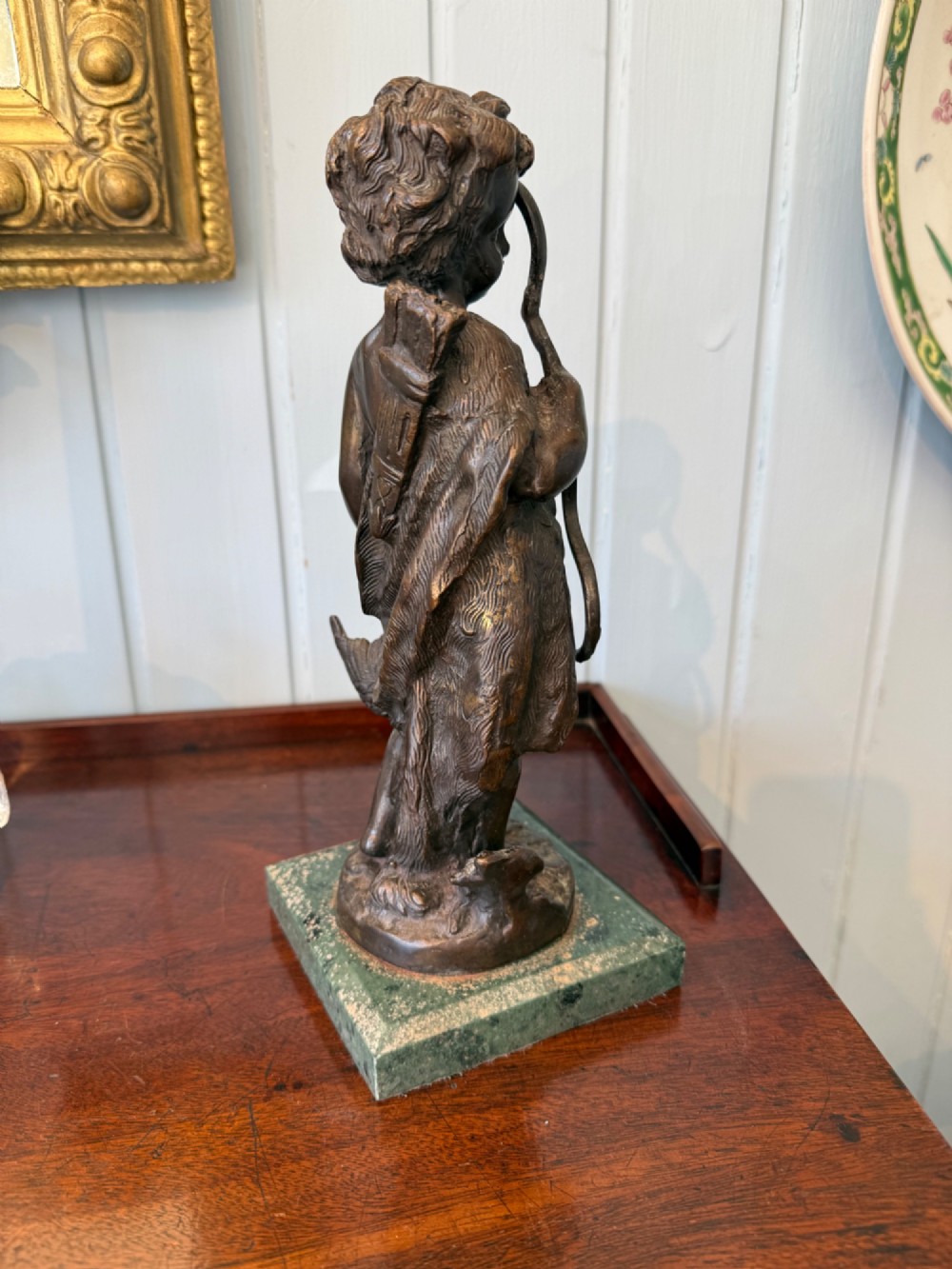An antique dark bronze figural sculpture depicting a young cherub holding a bow and hunted game, standing on a square green marble base. View IV