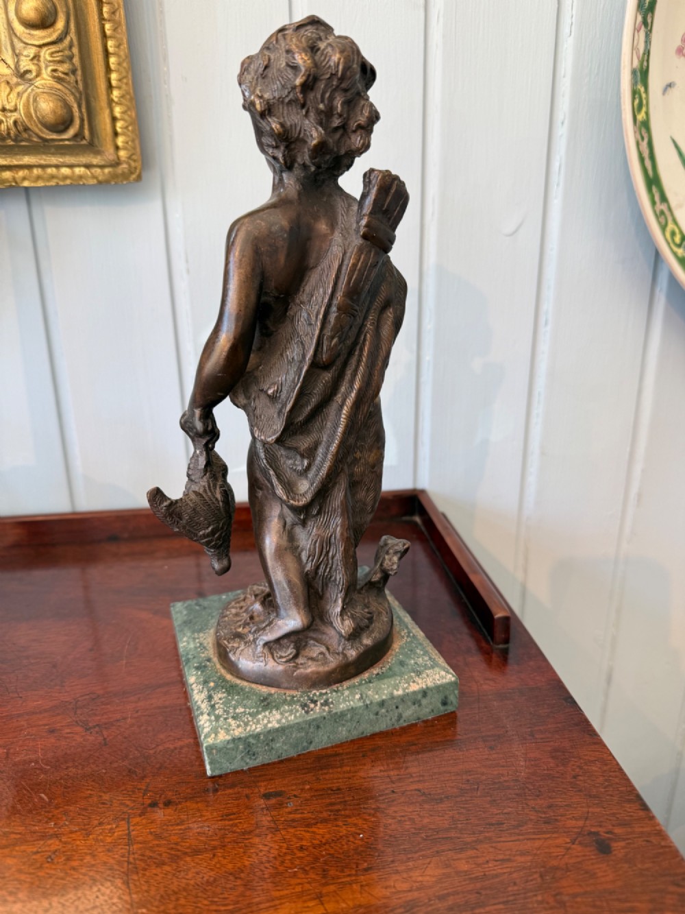 An antique dark bronze figural sculpture depicting a young cherub holding a bow and hunted game, standing on a square green marble base. View V