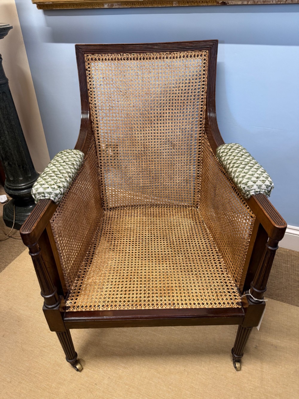 A late nineteenth-century dark mahogany Bergere armchair featuring woven cane side panels, fluted front legs on brass castors, and patterned upholstery. View IV