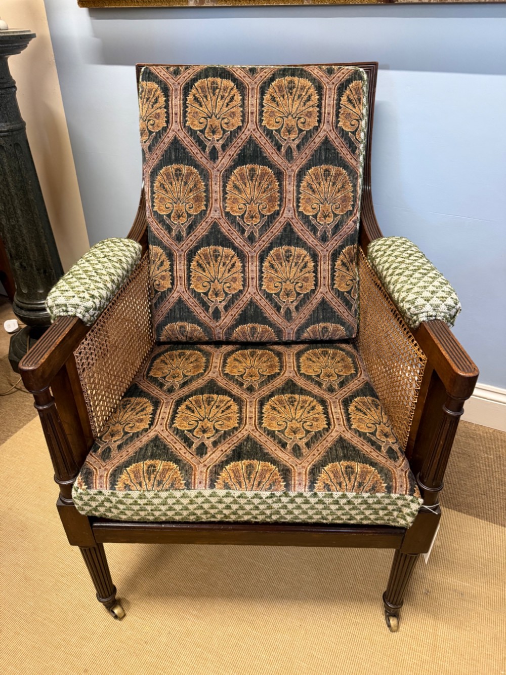 A late nineteenth-century dark mahogany Bergere armchair featuring woven cane side panels, fluted front legs on brass castors, and patterned upholstery. View V