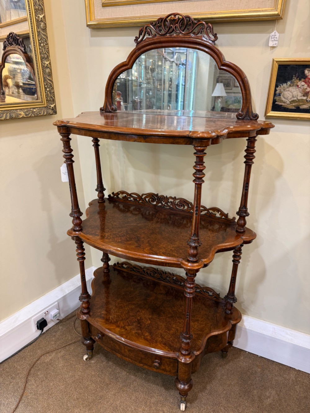 A three-tiered Victorian burr walnut whatnot featuring a carved fretwork upstand with a central arched mirror, turned spindle supports, and a base drawer on small castors.