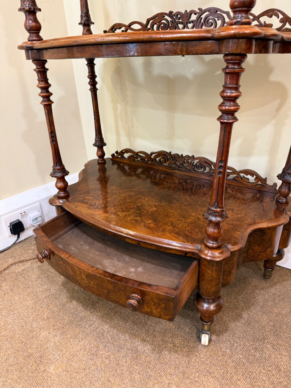 A three-tiered Victorian burr walnut whatnot featuring a carved fretwork upstand with a central arched mirror, turned spindle supports, and a base drawer on small castors. View IV