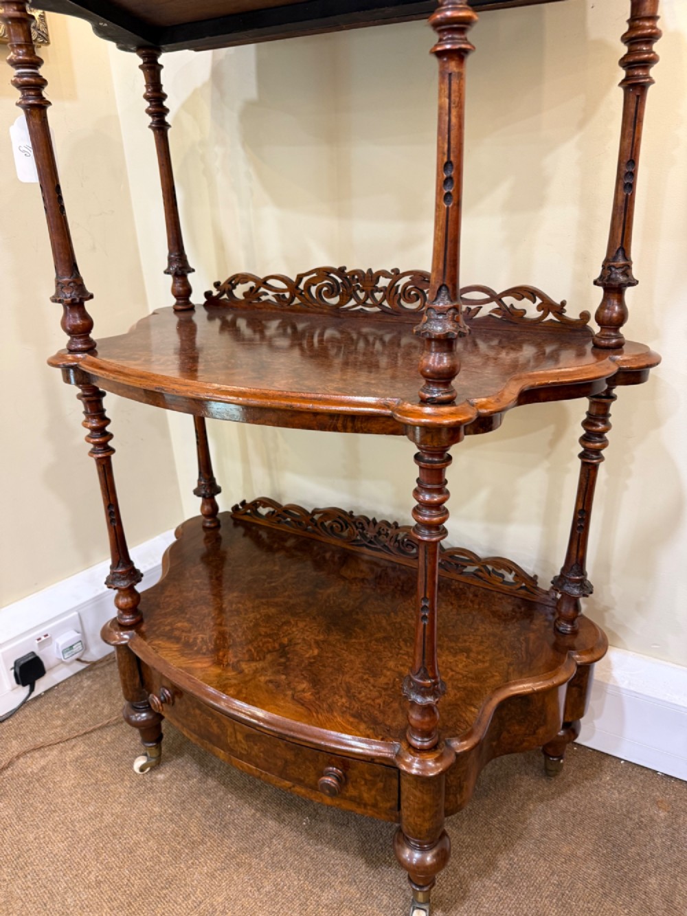 A three-tiered Victorian burr walnut whatnot featuring a carved fretwork upstand with a central arched mirror, turned spindle supports, and a base drawer on small castors. View V