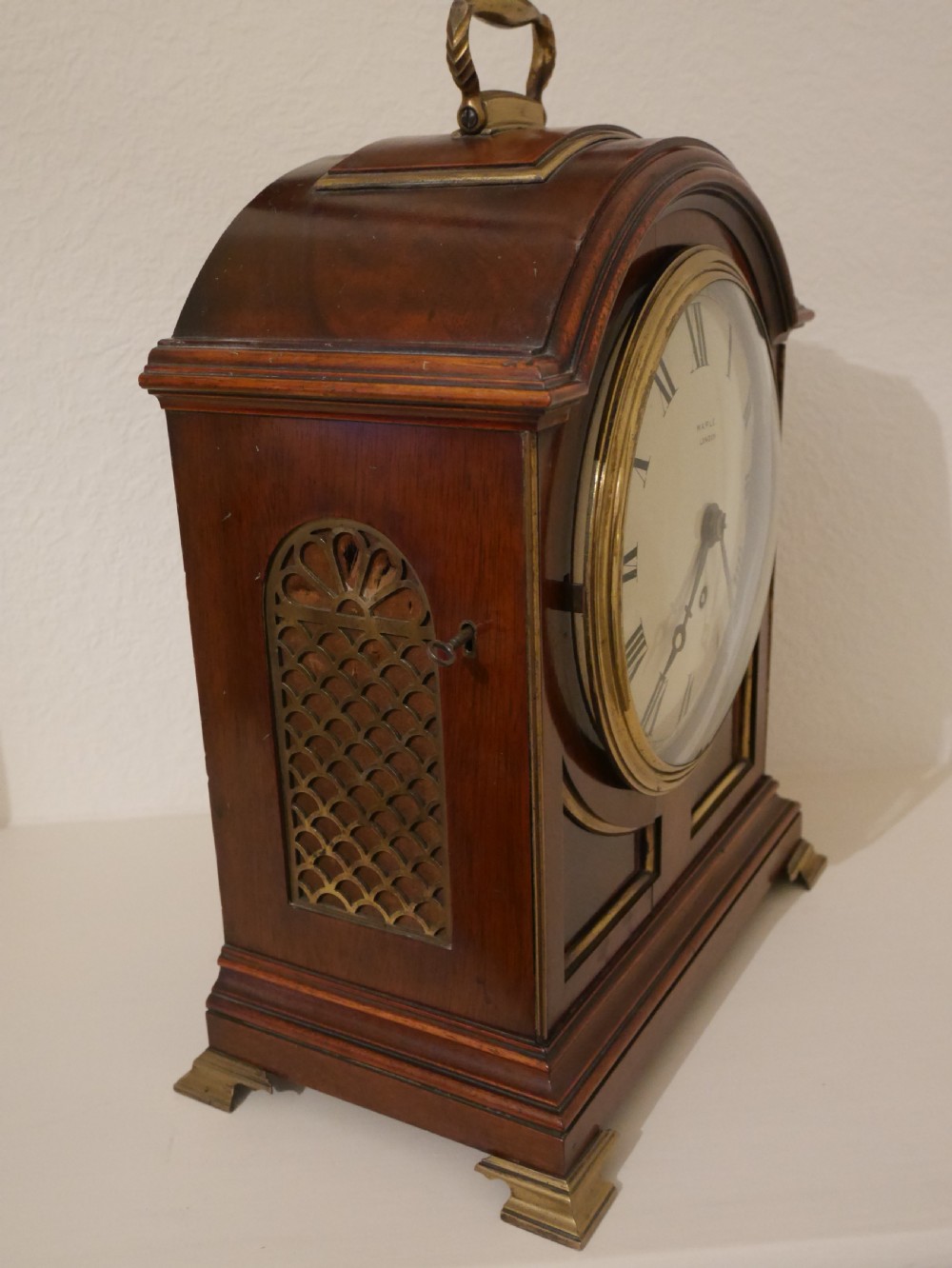 An antique mahogany bracket clock featuring an arched hood, top brass handle, brass bracket feet, and a white dial with Roman numerals. View V