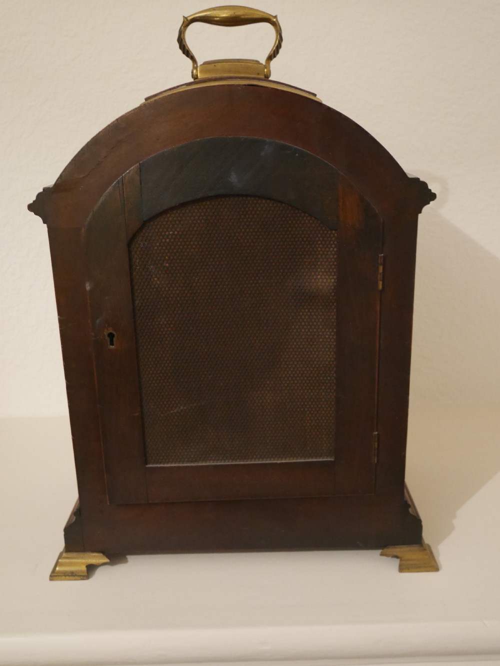 An antique mahogany bracket clock featuring an arched hood, top brass handle, brass bracket feet, and a white dial with Roman numerals. View VI