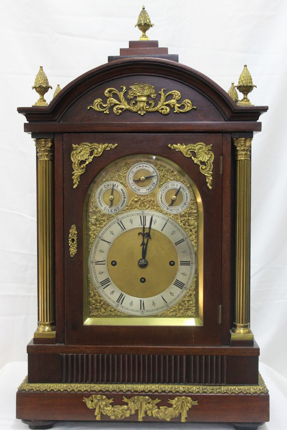 A dark mahogany Edwardian bracket clock featuring brass column supports, pineapple finials, and an ornate silvered dial with subsidiary chime indicators.