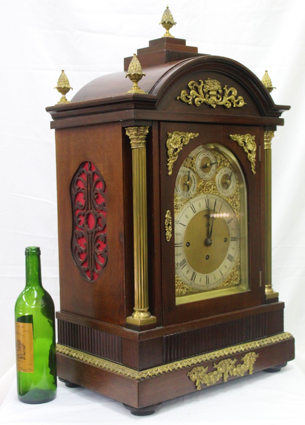 A dark mahogany Edwardian bracket clock featuring brass column supports, pineapple finials, and an ornate silvered dial with subsidiary chime indicators. View II