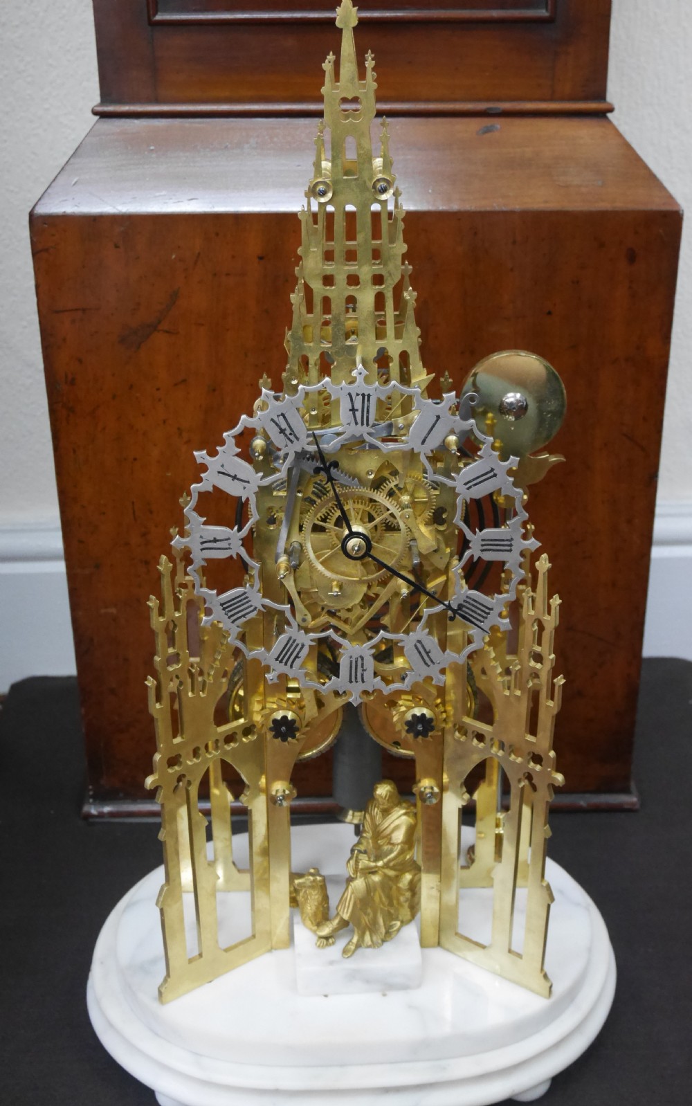 A Victorian brass skeleton clock designed in the shape of a Gothic spire, resting on a white marble base with a small seated figure at the bottom.