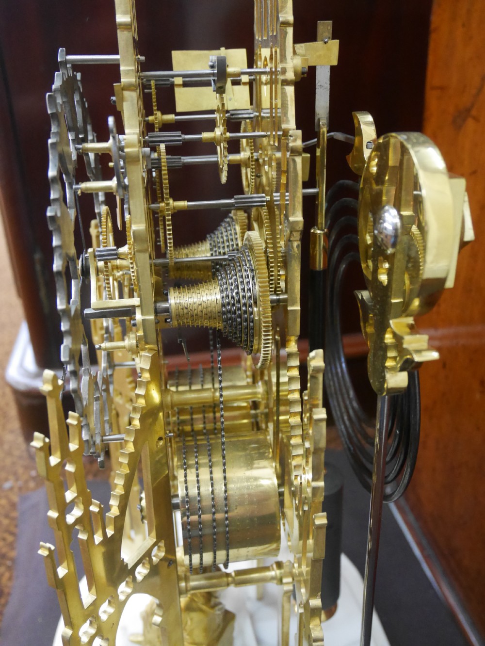 A Victorian brass skeleton clock designed in the shape of a Gothic spire, resting on a white marble base with a small seated figure at the bottom. View IV