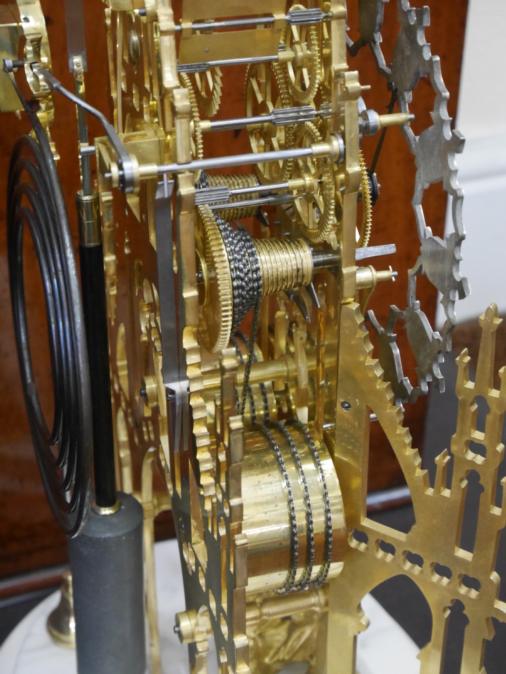 A Victorian brass skeleton clock designed in the shape of a Gothic spire, resting on a white marble base with a small seated figure at the bottom. View V