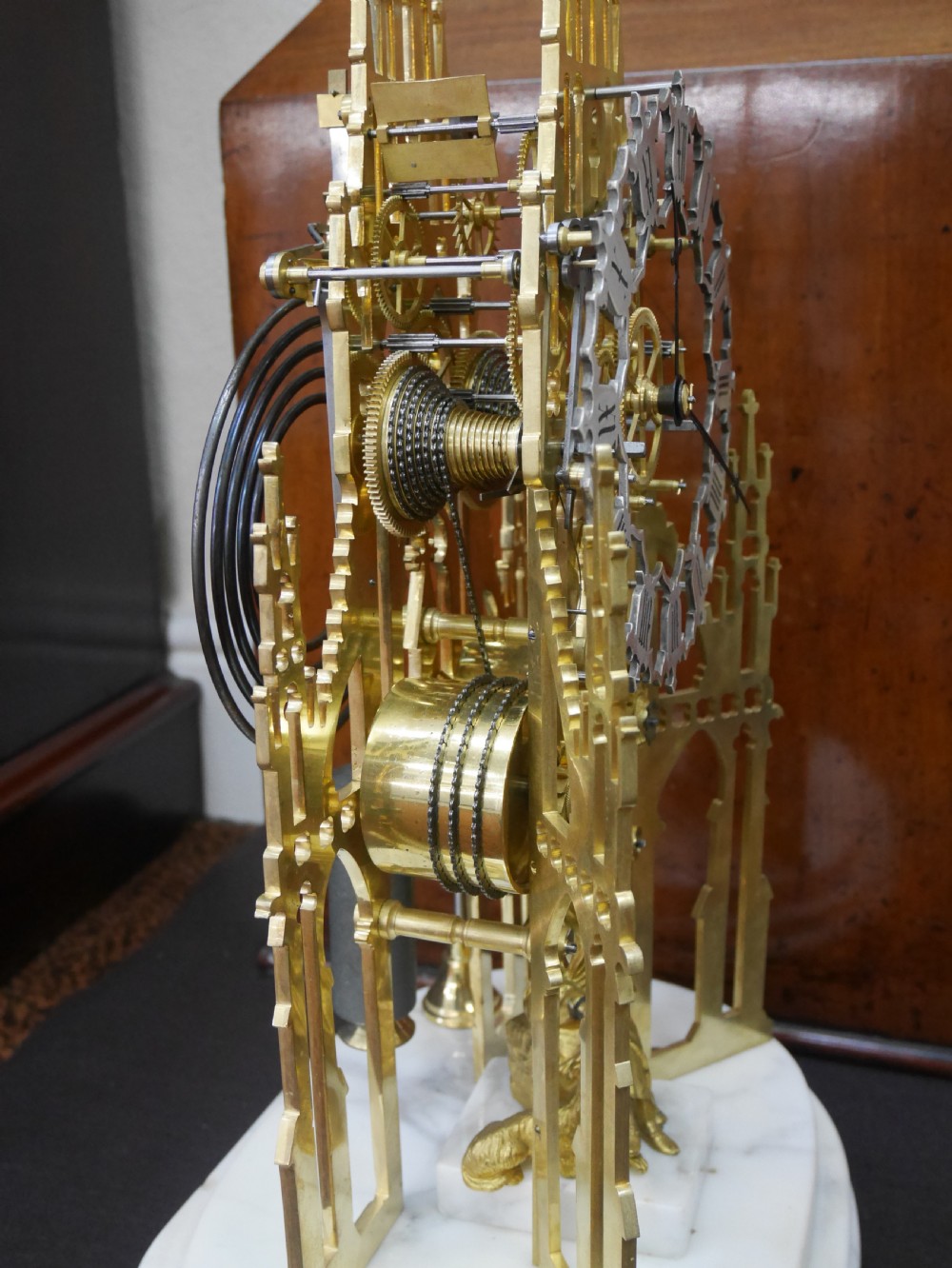A Victorian brass skeleton clock designed in the shape of a Gothic spire, resting on a white marble base with a small seated figure at the bottom. View VI
