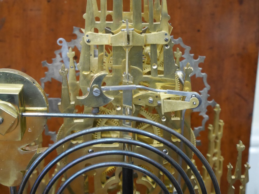 A Victorian brass skeleton clock designed in the shape of a Gothic spire, resting on a white marble base with a small seated figure at the bottom. View VIII
