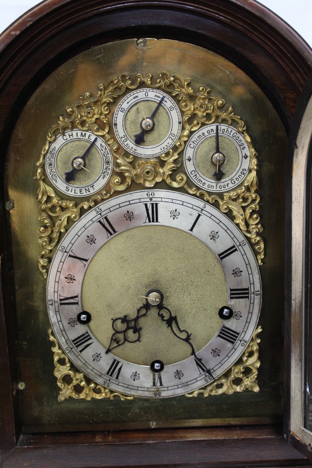 An arched mahogany bracket clock featuring brass spandrels, subsidiary dials, a silvered chapter ring, and brass bracket feet. View VII