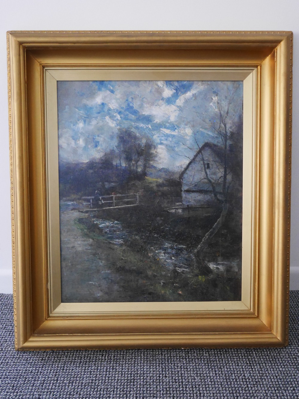 A framed oil painting depicting a rural cottage alongside a small stream beneath a cloudy blue sky, housed in a wide gilt frame.