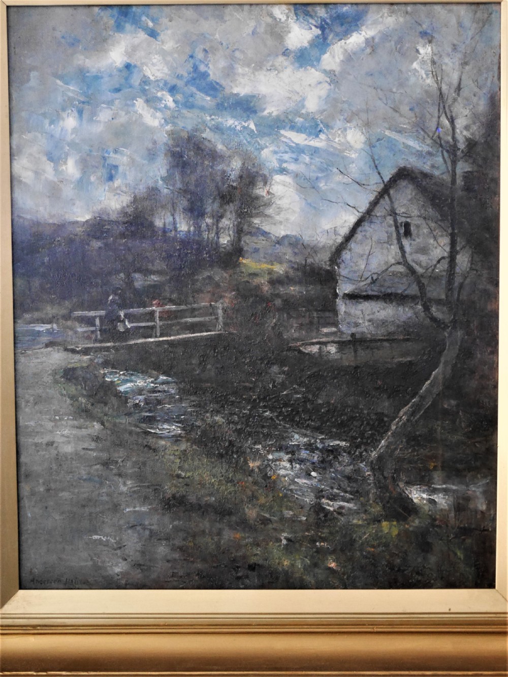 A framed oil painting depicting a rural cottage alongside a small stream beneath a cloudy blue sky, housed in a wide gilt frame. View II