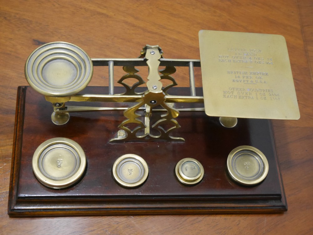 Victorian brass letter scales with engraved postal rates mounted on a dark mahogany base beside four nested brass weights.