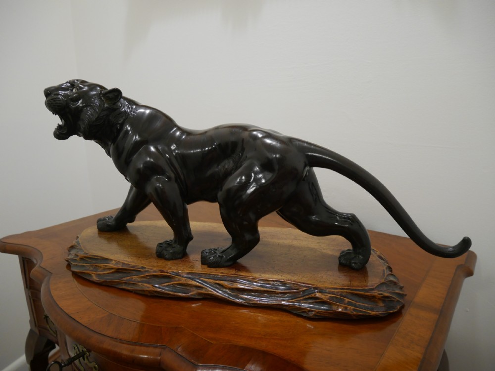 A dark bronze Japanese tiger sculpture depicted in a roaring, prowling stance mounted on a carved wooden base.