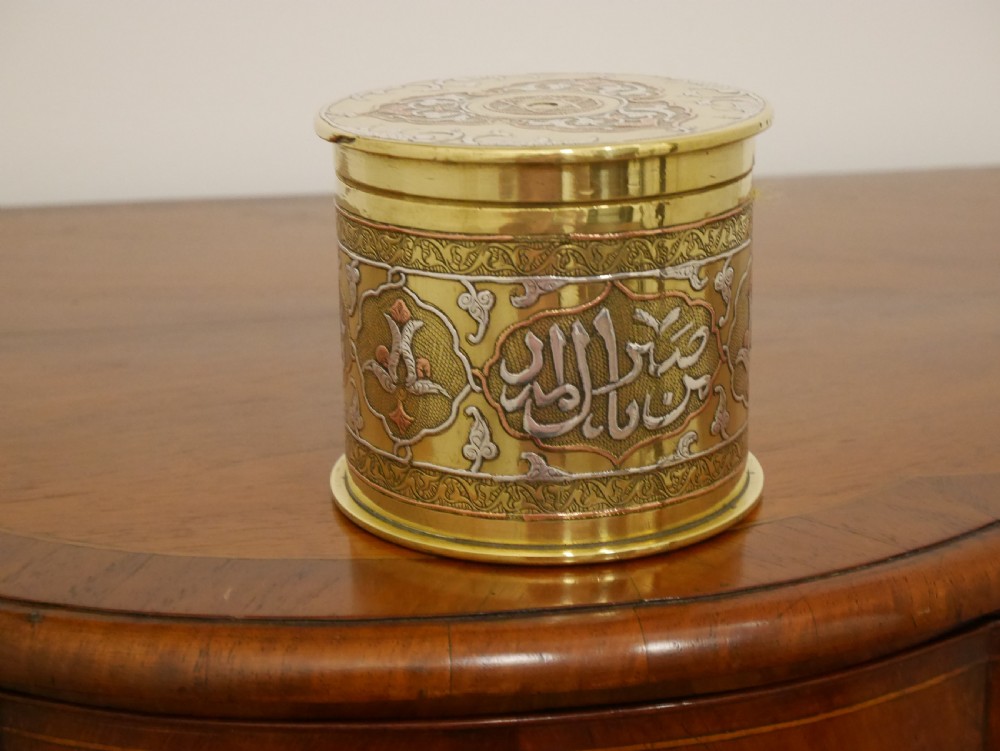 A cylindrical brass artillery shell casing converted into a lidded container with silver and copper Middle Eastern calligraphy inlays.