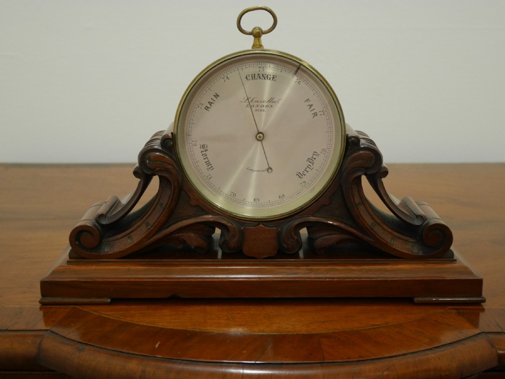 A mid-Victorian aneroid desk barometer mounted in a carved mahogany case on a stepped base with a brass dial bezel.