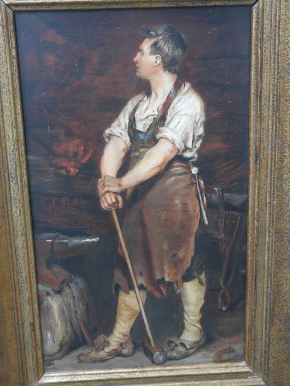 A framed oil painting depicting a blacksmith standing in profile inside a forge, holding a sledgehammer while wearing an apron, presented in a wide gilded frame. View III