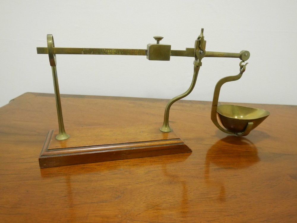 An antique brass balance scale featuring a graduated measurement beam, sliding weight, and single suspended pan mounted on a mahogany base.