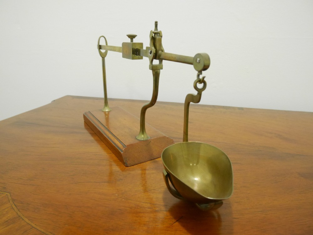 An antique brass balance scale featuring a graduated measurement beam, sliding weight, and single suspended pan mounted on a mahogany base. View IV