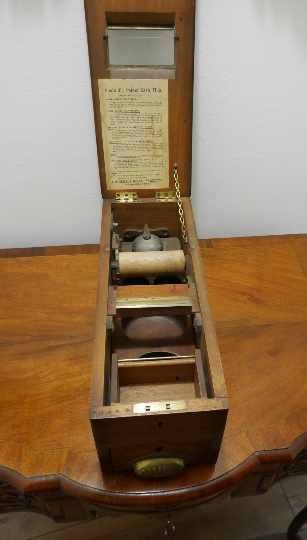 An open mahogany Gledhill patent cash till showing internal mechanical brass parts, a paper roll mechanism, and a printed paper instruction label on the underside of the lid.