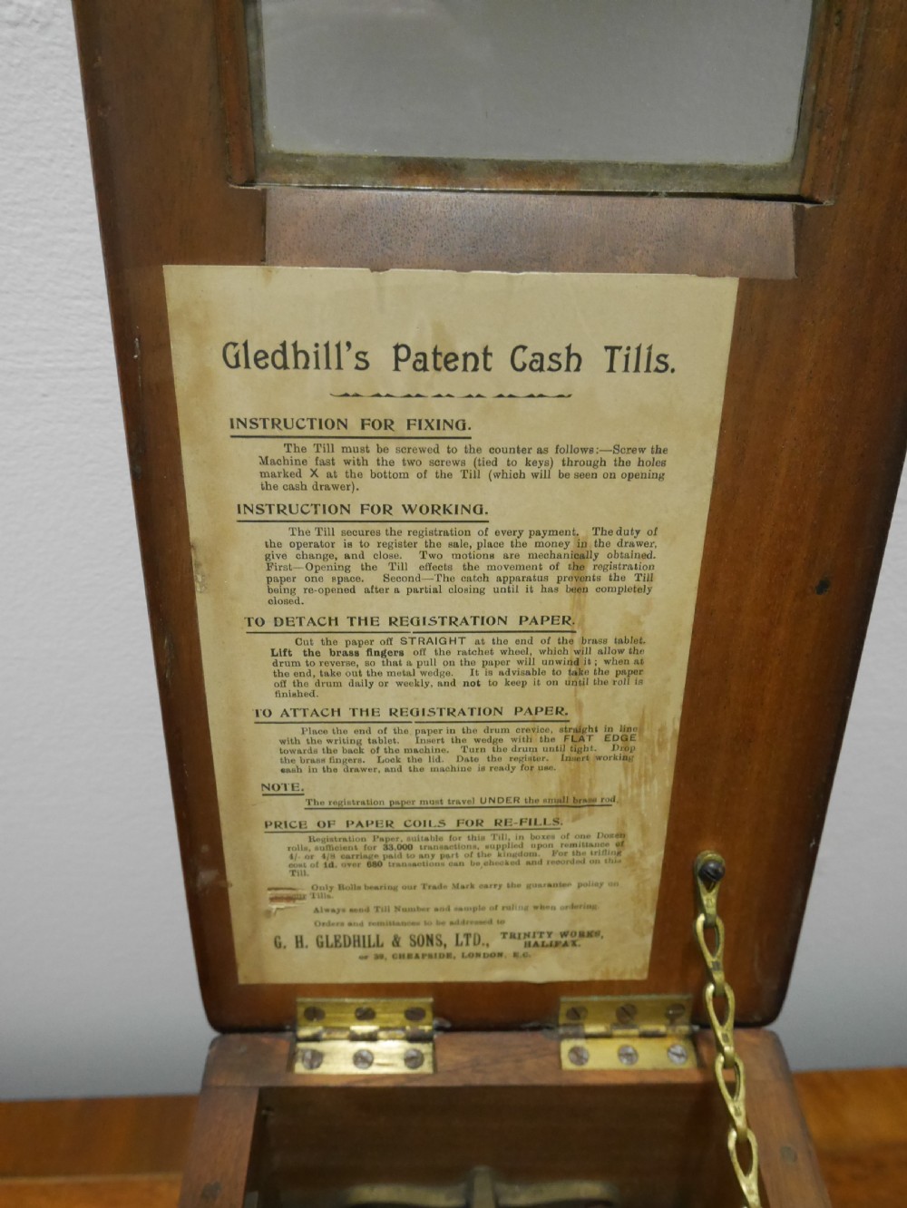 An open mahogany Gledhill patent cash till showing internal mechanical brass parts, a paper roll mechanism, and a printed paper instruction label on the underside of the lid. View V