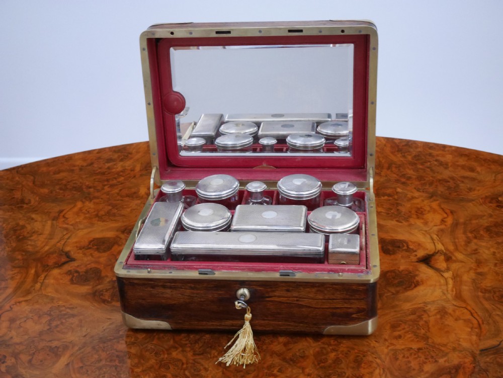 An open antique brass-bound wooden dressing box featuring a red velvet interior, integrated lid mirror, and fitted glass bottles with metal caps.