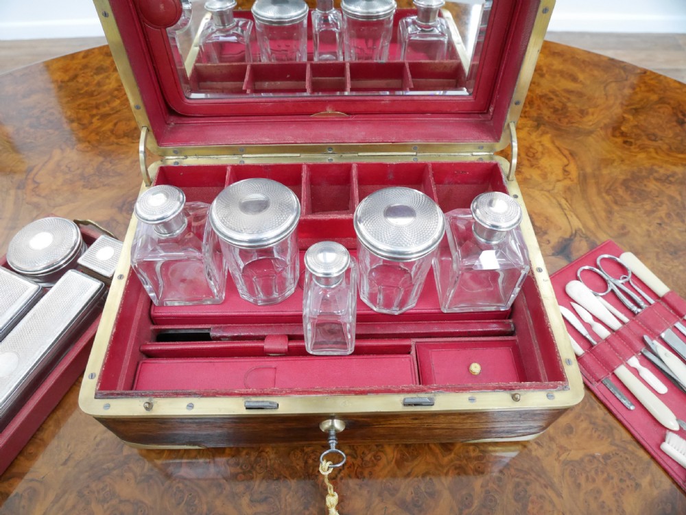 An open antique brass-bound wooden dressing box featuring a red velvet interior, integrated lid mirror, and fitted glass bottles with metal caps. View II