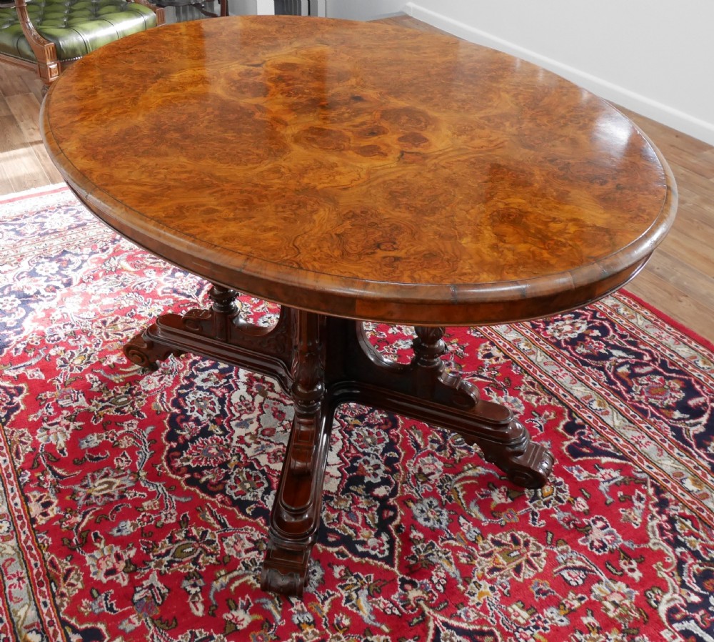 An oval Victorian burr walnut tilt-top loo table featuring figured timber grain and a carved central pedestal base with four scroll feet.