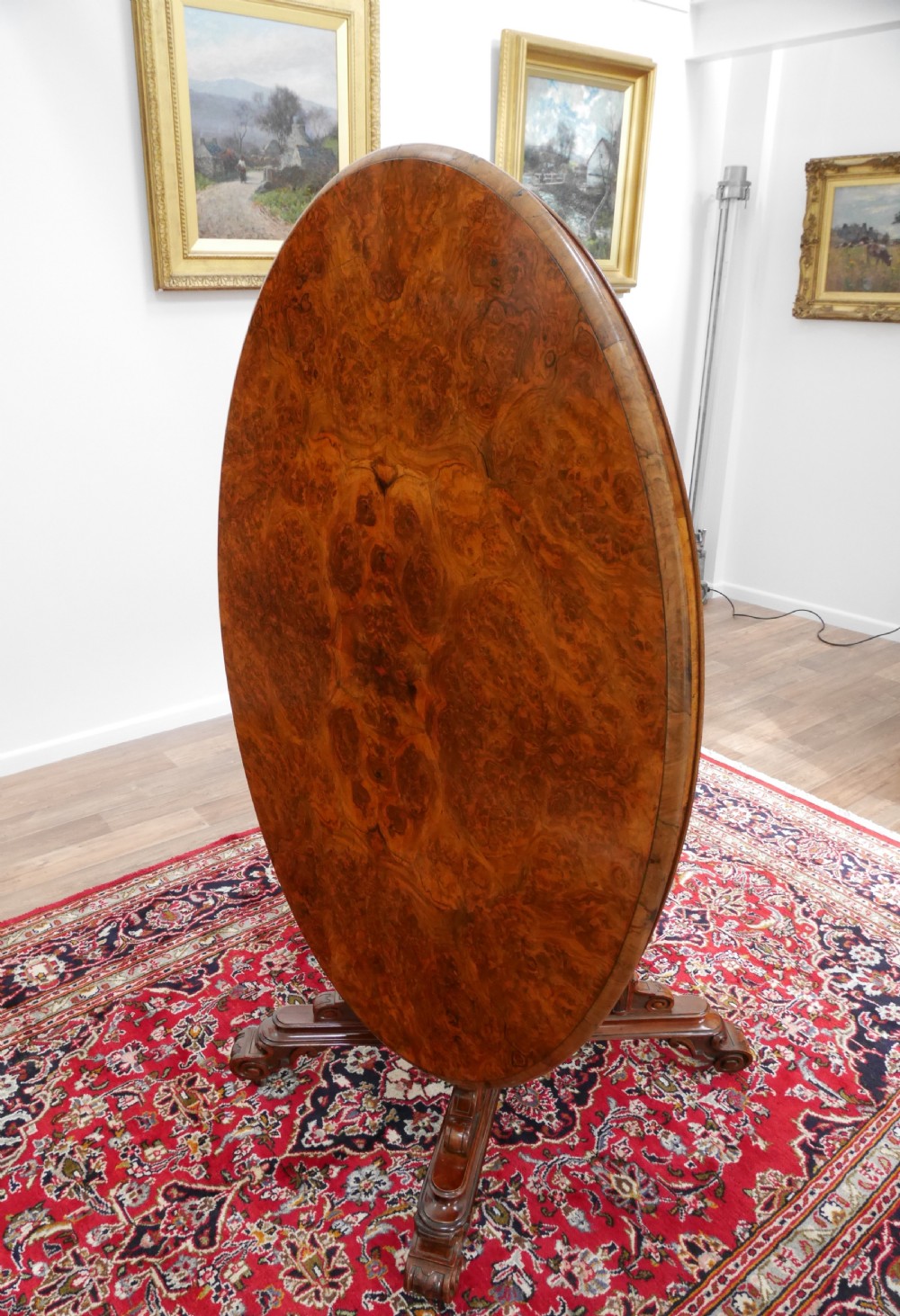 An oval Victorian burr walnut tilt-top loo table featuring figured timber grain and a carved central pedestal base with four scroll feet. View II