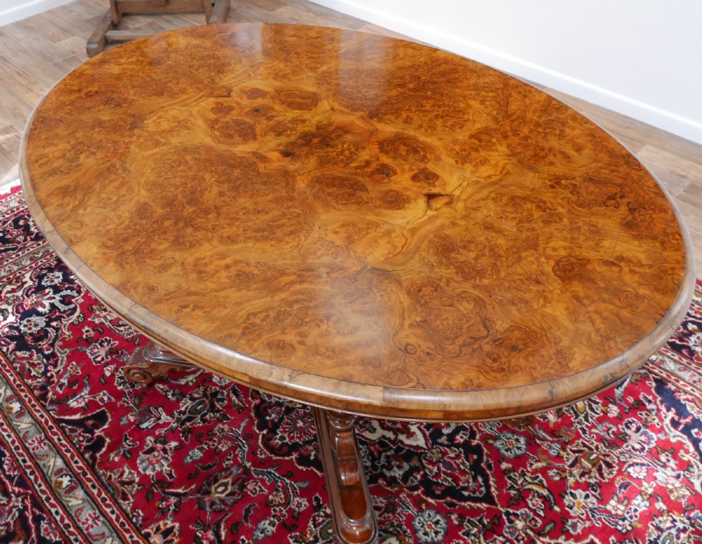 An oval Victorian burr walnut tilt-top loo table featuring figured timber grain and a carved central pedestal base with four scroll feet. View IV