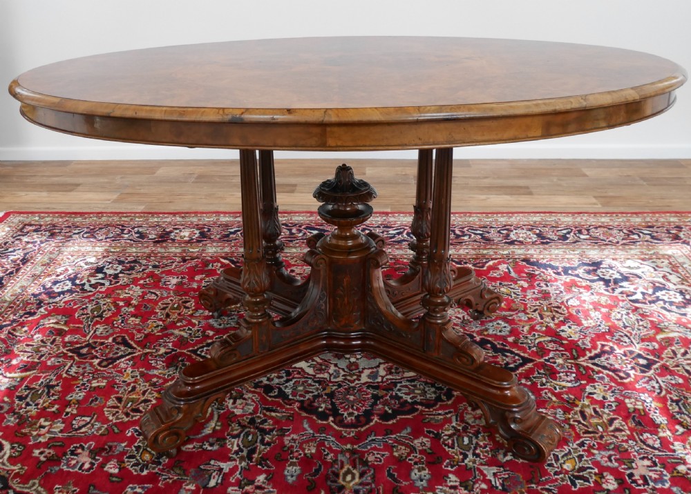 An oval Victorian burr walnut tilt-top loo table featuring figured timber grain and a carved central pedestal base with four scroll feet. View V