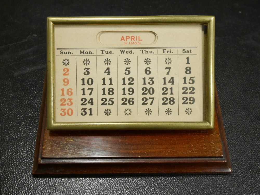 A vintage perpetual desk calendar with a polished brass frame holding paper month cards, mounted on a dark mahogany wooden base.