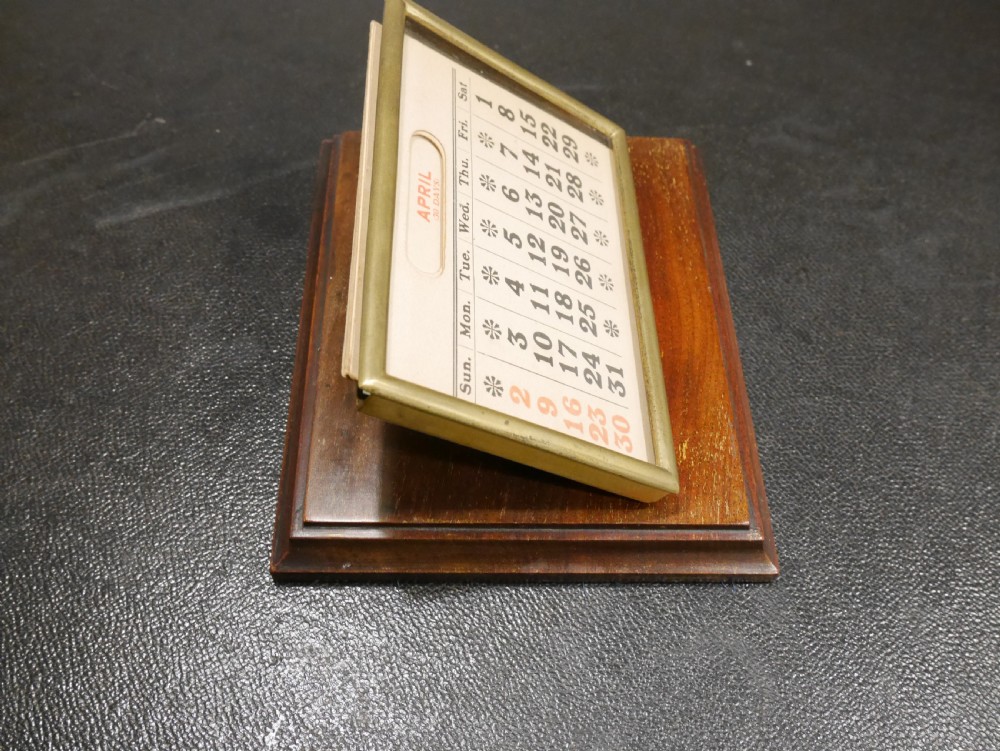 A vintage perpetual desk calendar with a polished brass frame holding paper month cards, mounted on a dark mahogany wooden base. View II