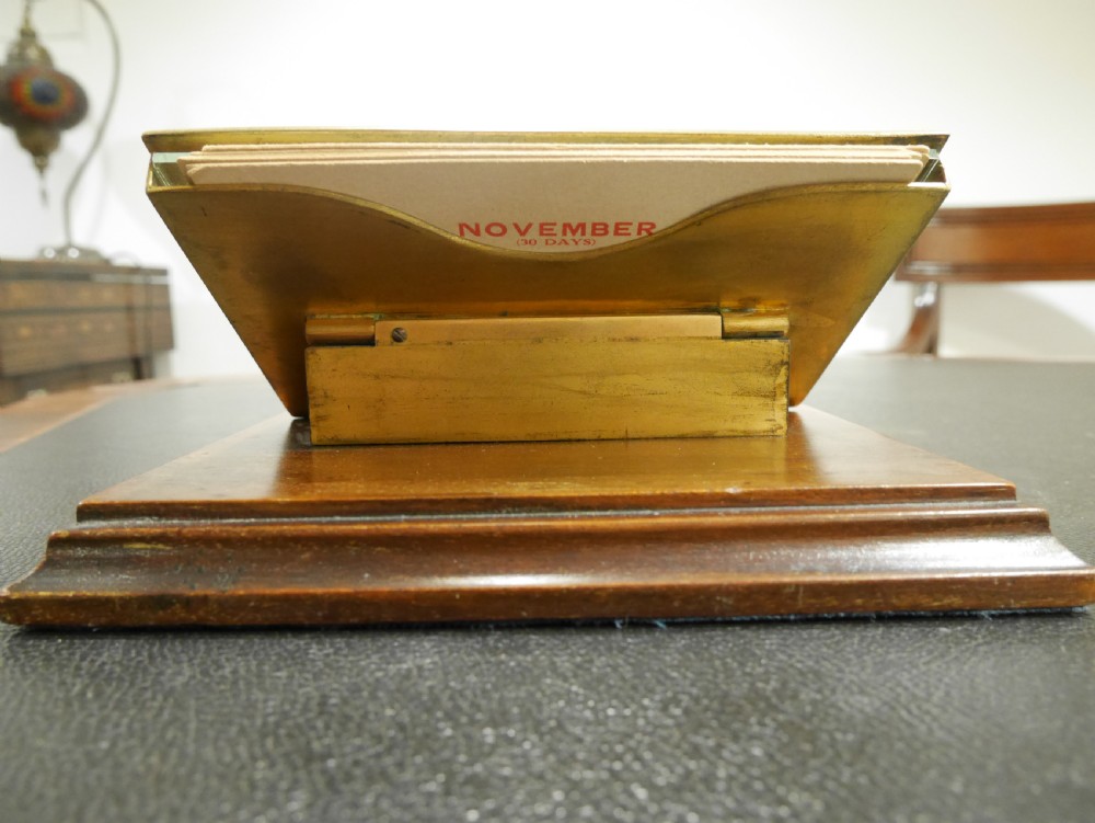 A vintage perpetual desk calendar with a polished brass frame holding paper month cards, mounted on a dark mahogany wooden base. View III