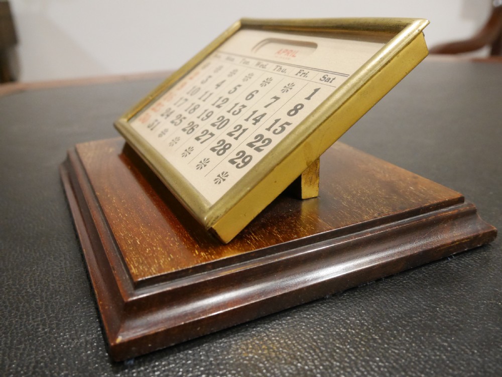 A vintage perpetual desk calendar with a polished brass frame holding paper month cards, mounted on a dark mahogany wooden base. View IV